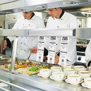 Food service line with chefs in white uniforms and hats standing behind glass partition. Various food items and plates are visible on the counter.